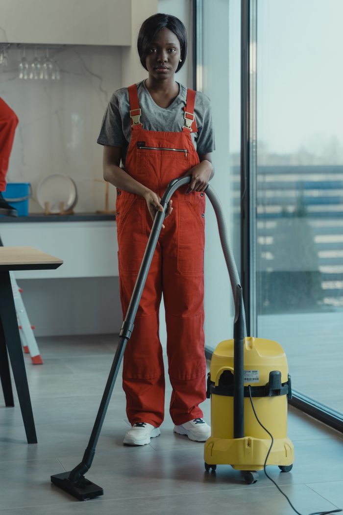 creative-03 An adult woman wearing red coveralls is using a vacuum cleaner in a contemporary indoor space, with her attention centered on cleaning work.